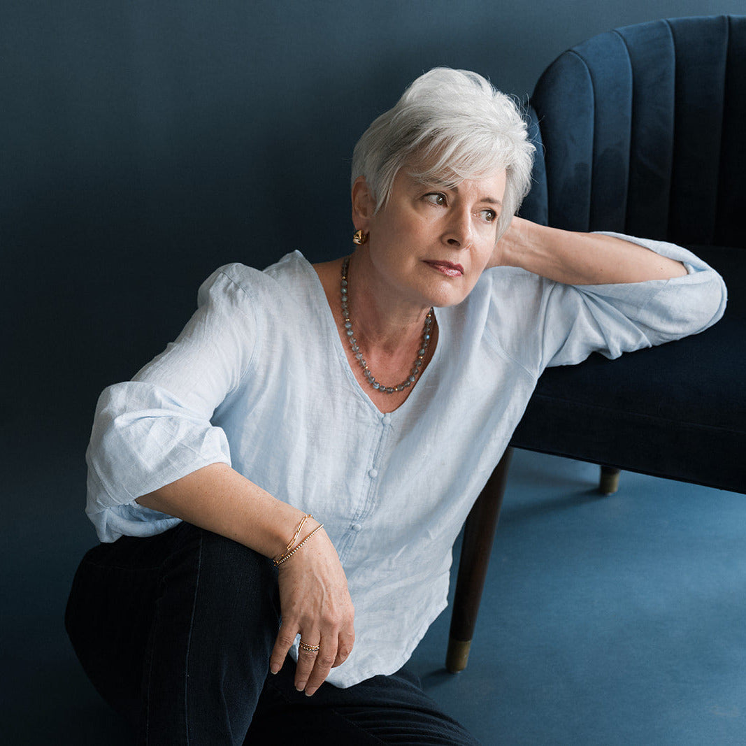 Woman wearing round labradorite gem necklace with gold beads in light blue shirt leaning on a blue chair