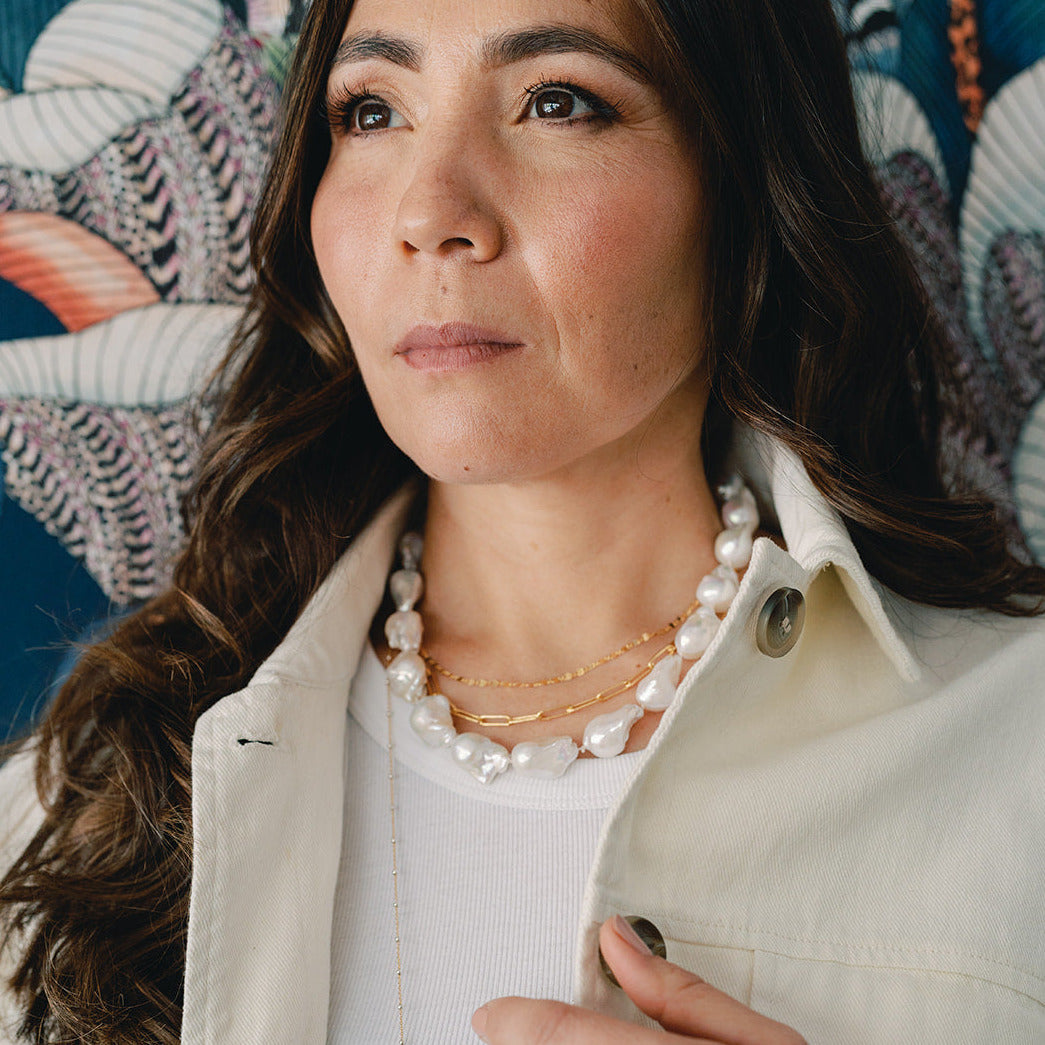 Woman on patterned background in white jacket and large pearl necklace