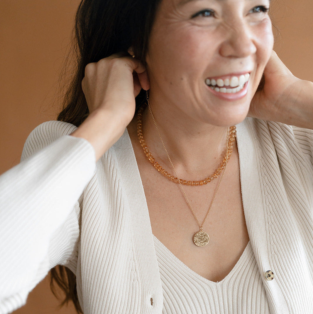 Woman smiling and putting on a gold pendant, layered with a topaz necklace