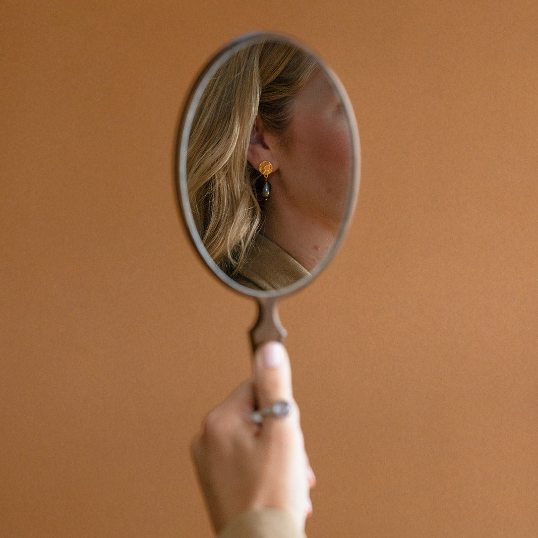 Woman holding mirror showing reflection of her wearing a peacock pearl earring