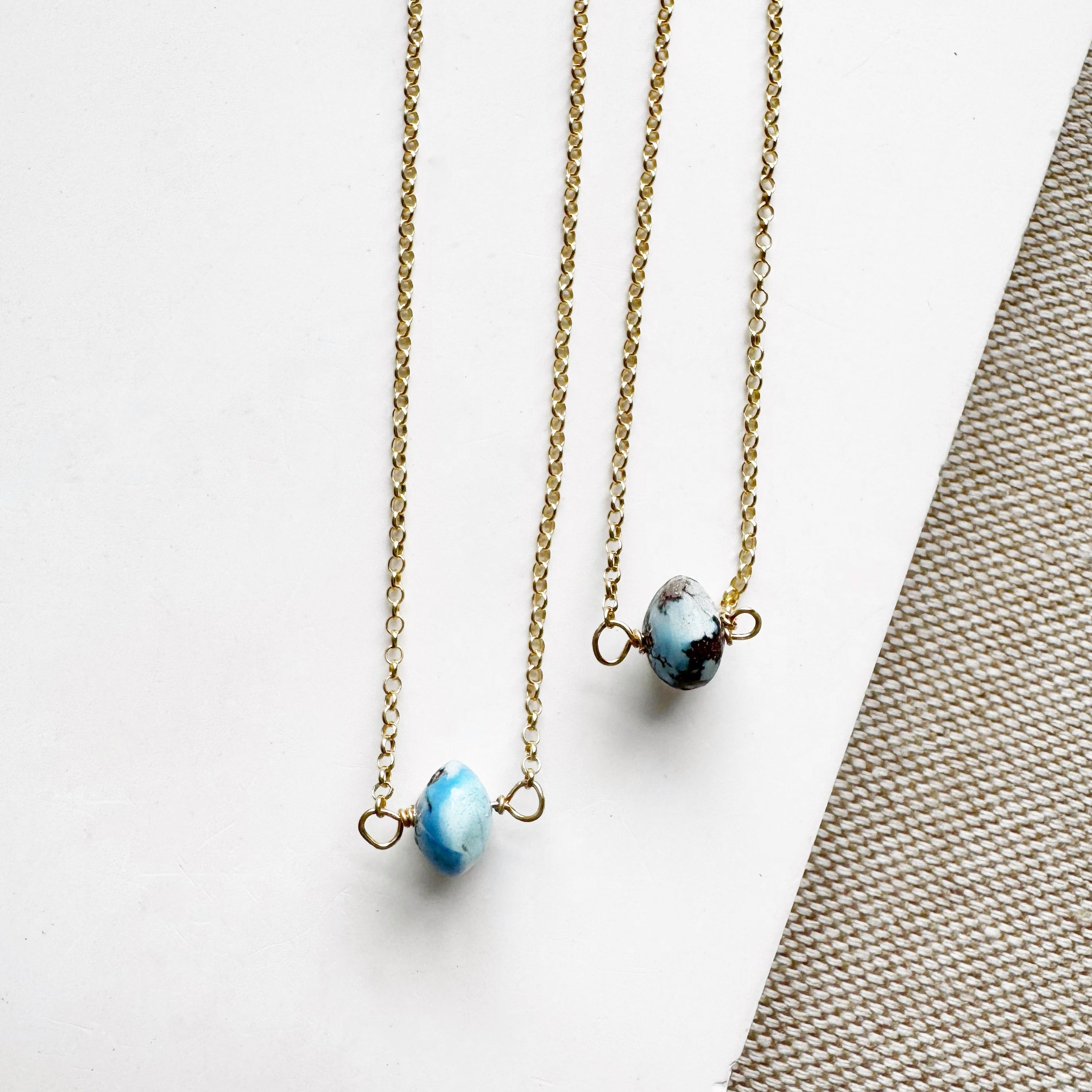 Two necklaces with a single small Golden Hill turquoise bead on gold chains on a white surface.