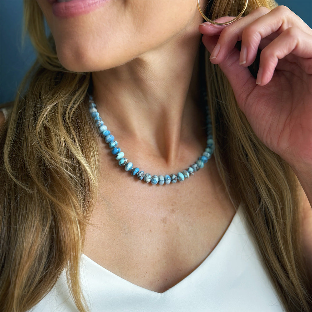 A close-up lifestyle shot of a woman wearing a chunky beaded necklace made of light blue and white marbled beads, paired with a white top and gold hoop earrings.