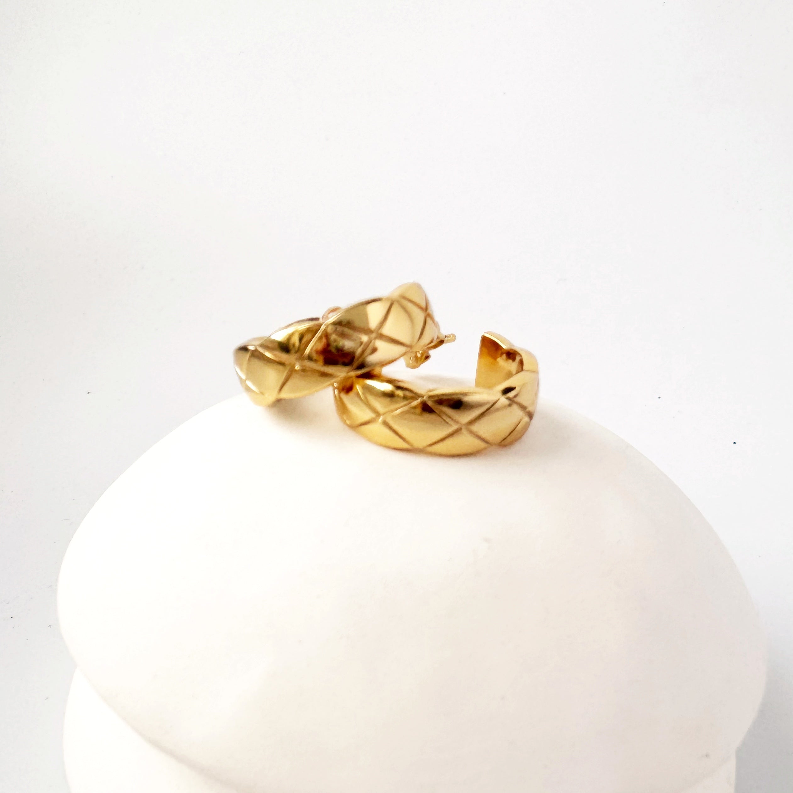 A pair of gold hoop earrings with a quilted design, displayed on a white stand.