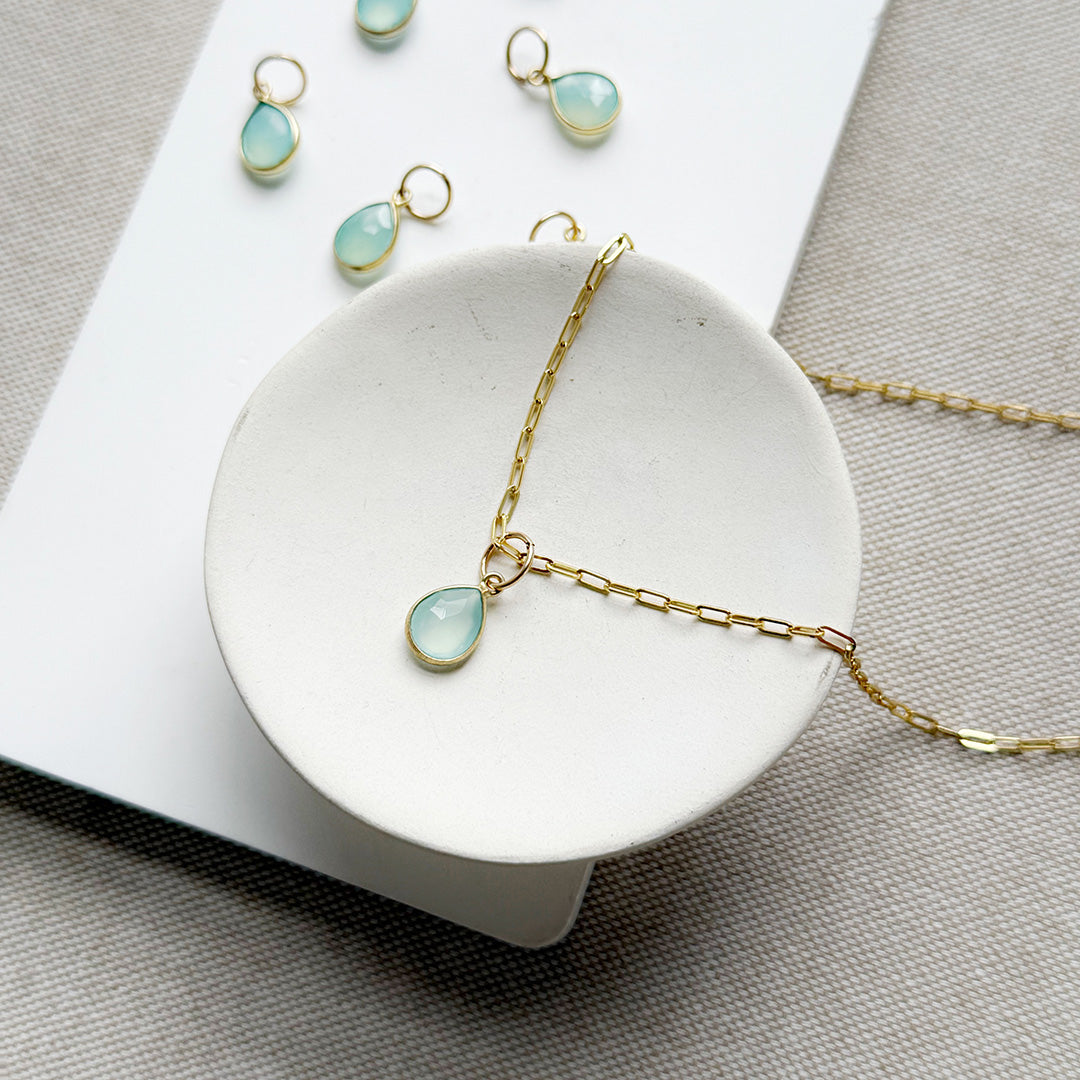 Teardrop shaped aqua chalcedony charm on a gold chain in a white dish