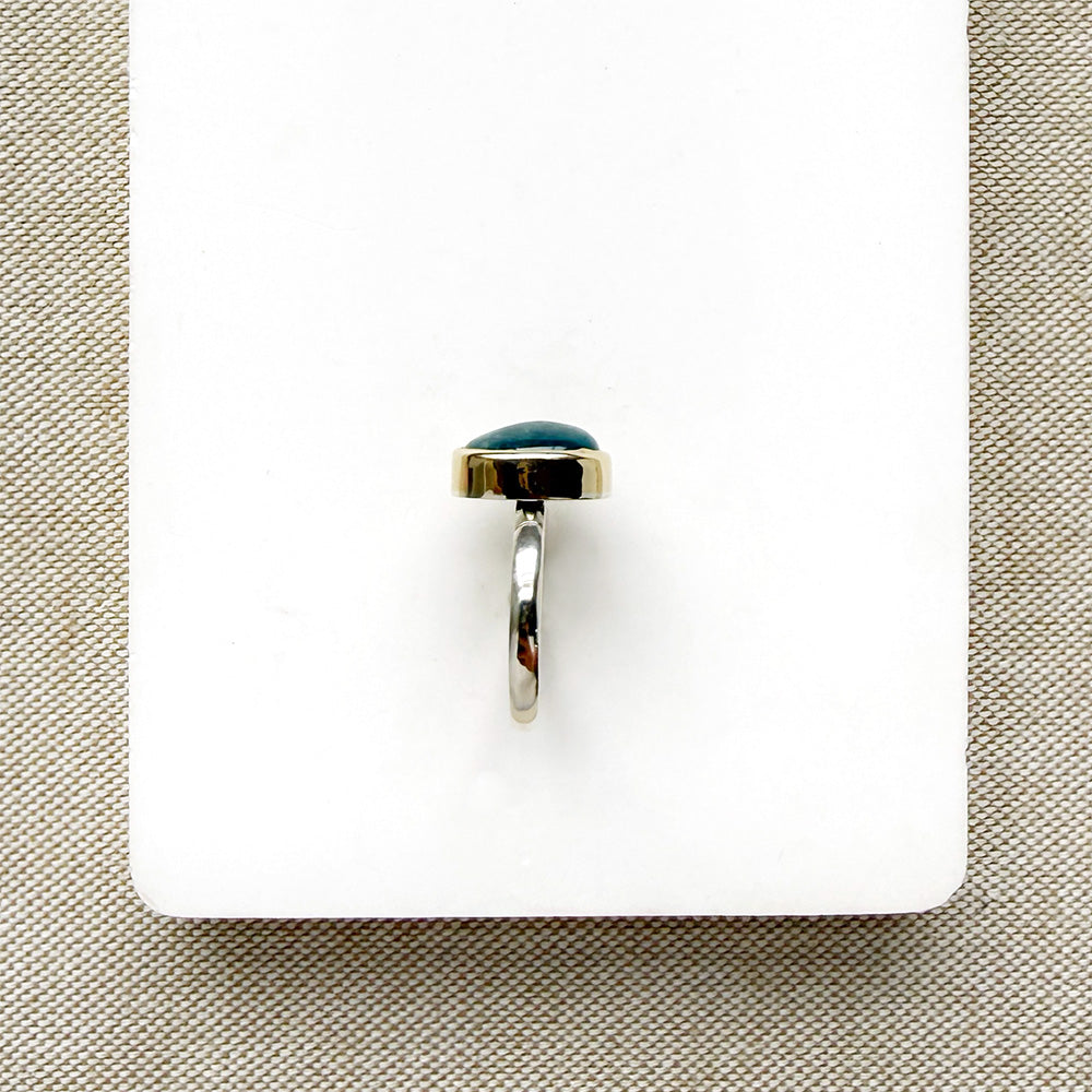 A silver ring with a small oval aquamarine cabochon gemstone, displayed on a white rectangular card over a beige woven fabric background.