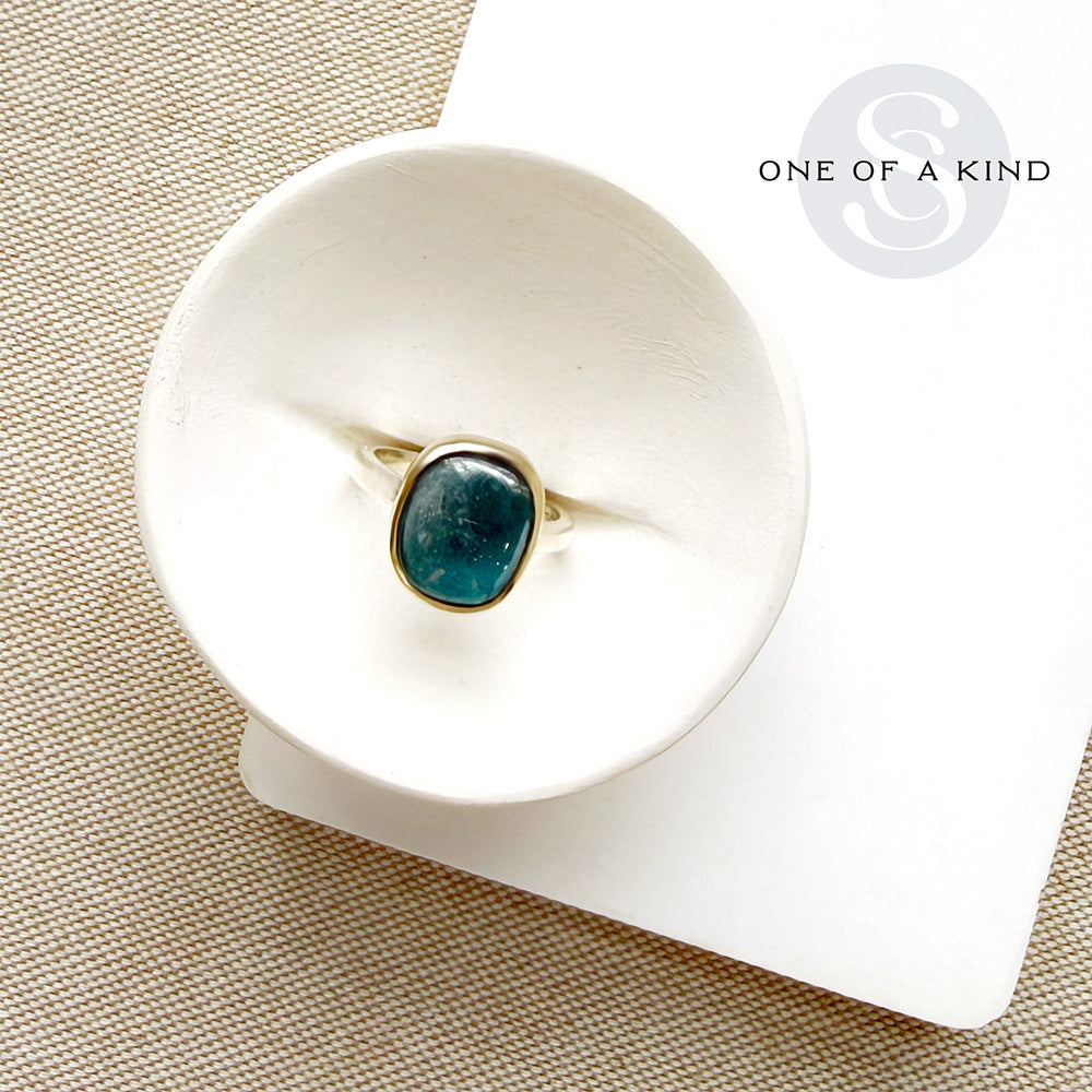 A gold ring with a round teal-blue stone sits in a white ceramic dish. The dish is placed on a textured beige fabric surface next to a white card with the text 'ONE OF A KIND'.