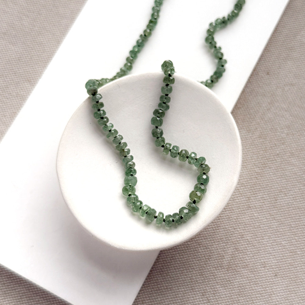A beaded gemstone necklace made of green kyanite with a gold-tone clasp and extender chain, displayed in a white dish