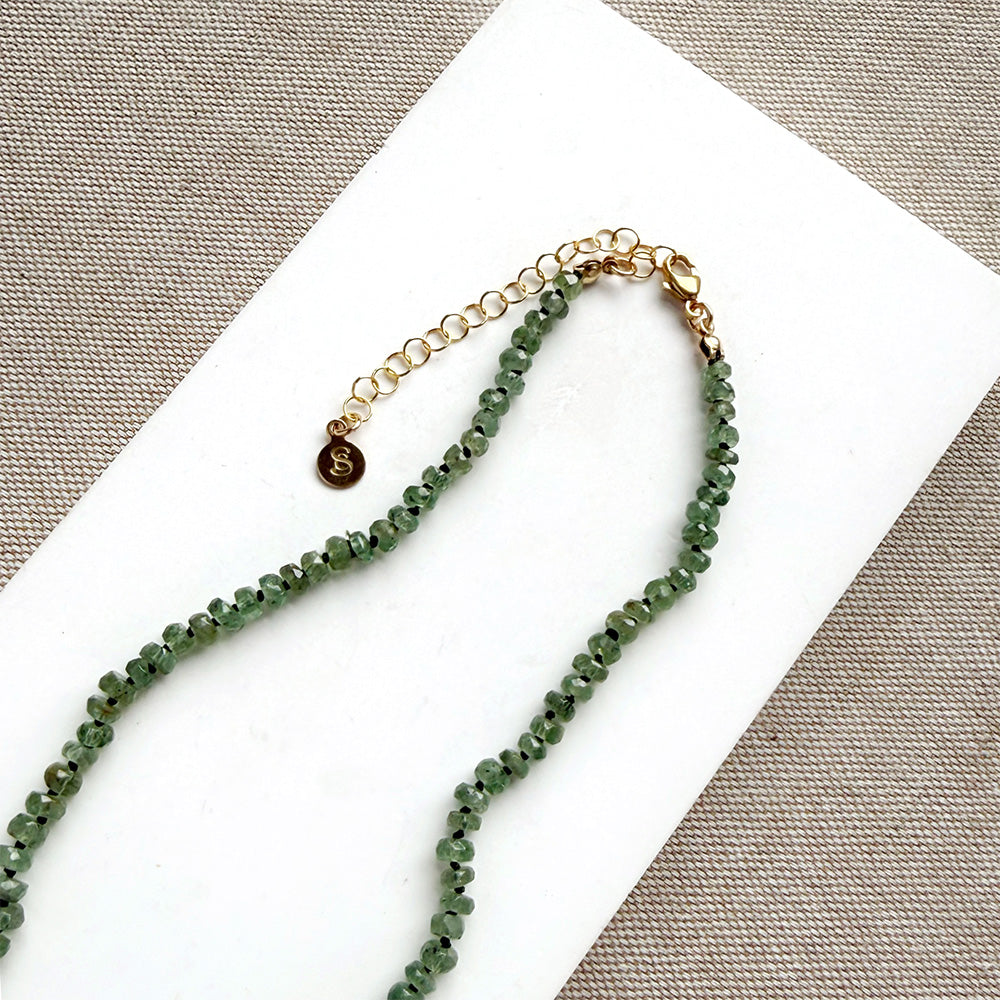 A beaded gemstone necklace made of green kyanite with a gold-tone clasp and extender chain, displayed on a white rectangular card against a textured gray fabric background.