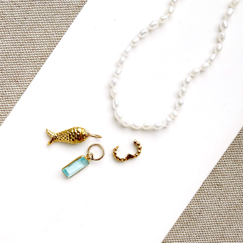 A white pearl necklace with a gold fish charm, a small gold ring, and a light blue rectangular charm, arranged on a white card with textured beige fabric in the corners.