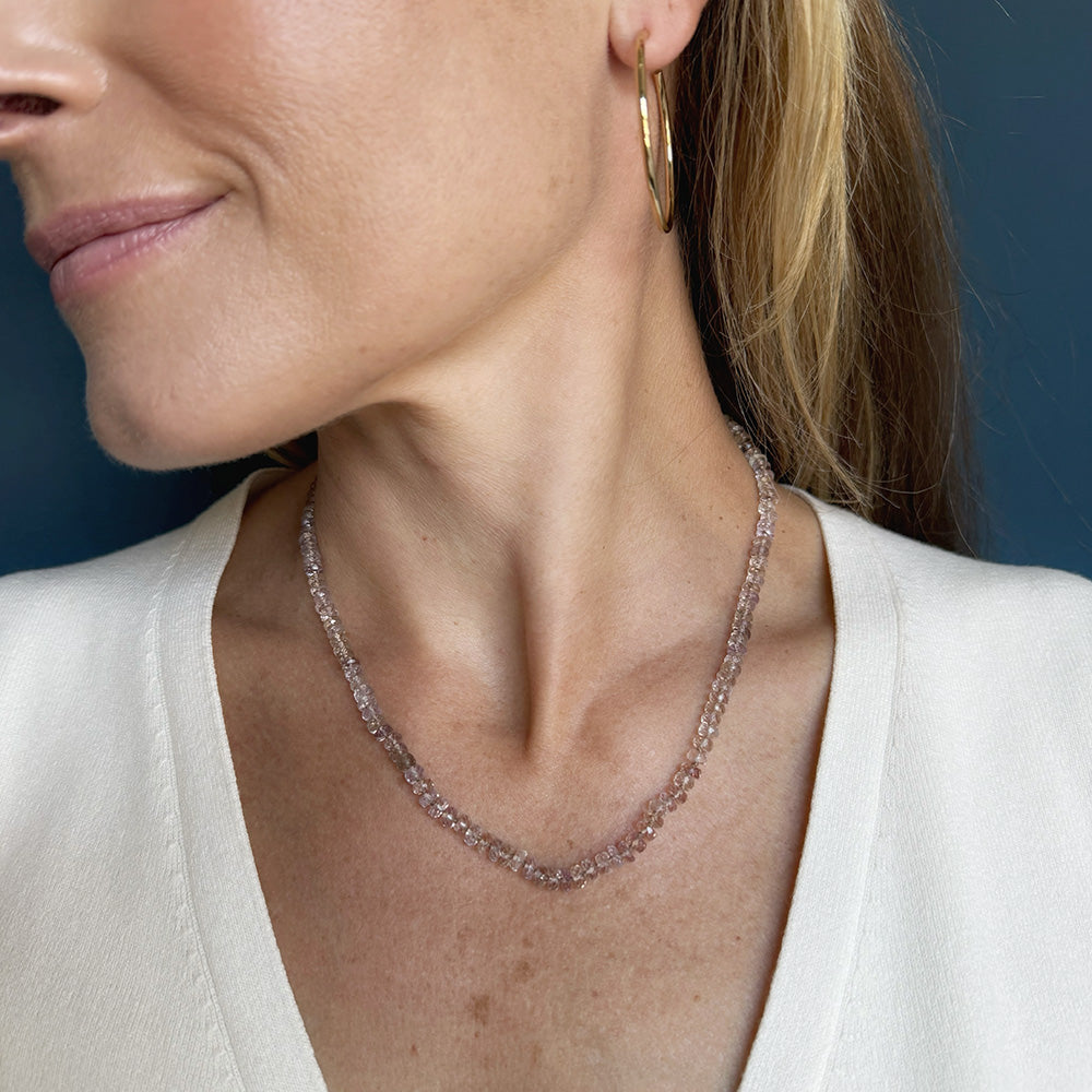 A close-up lifestyle shot of a woman wearing a delicate rose gold chain necklace and a gold hoop earring against a dark blue background.
