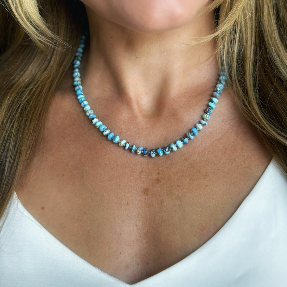 A close-up of a woman wearing a knotted Golden Hills turquoise necklace, paired with a white top against a dark blue background.