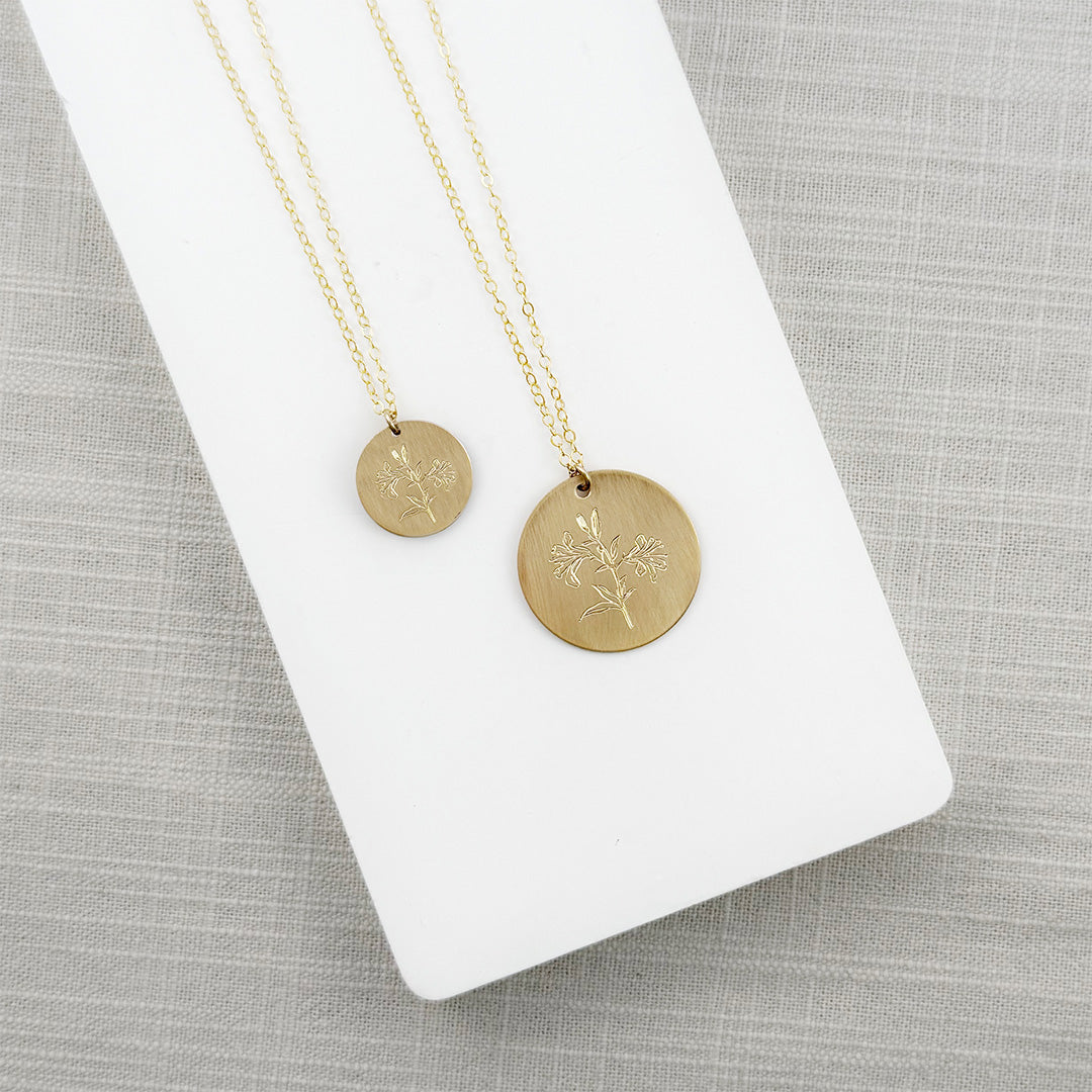 Two gold necklaces with round pendants, one small and one large, on a white stand against a gray background