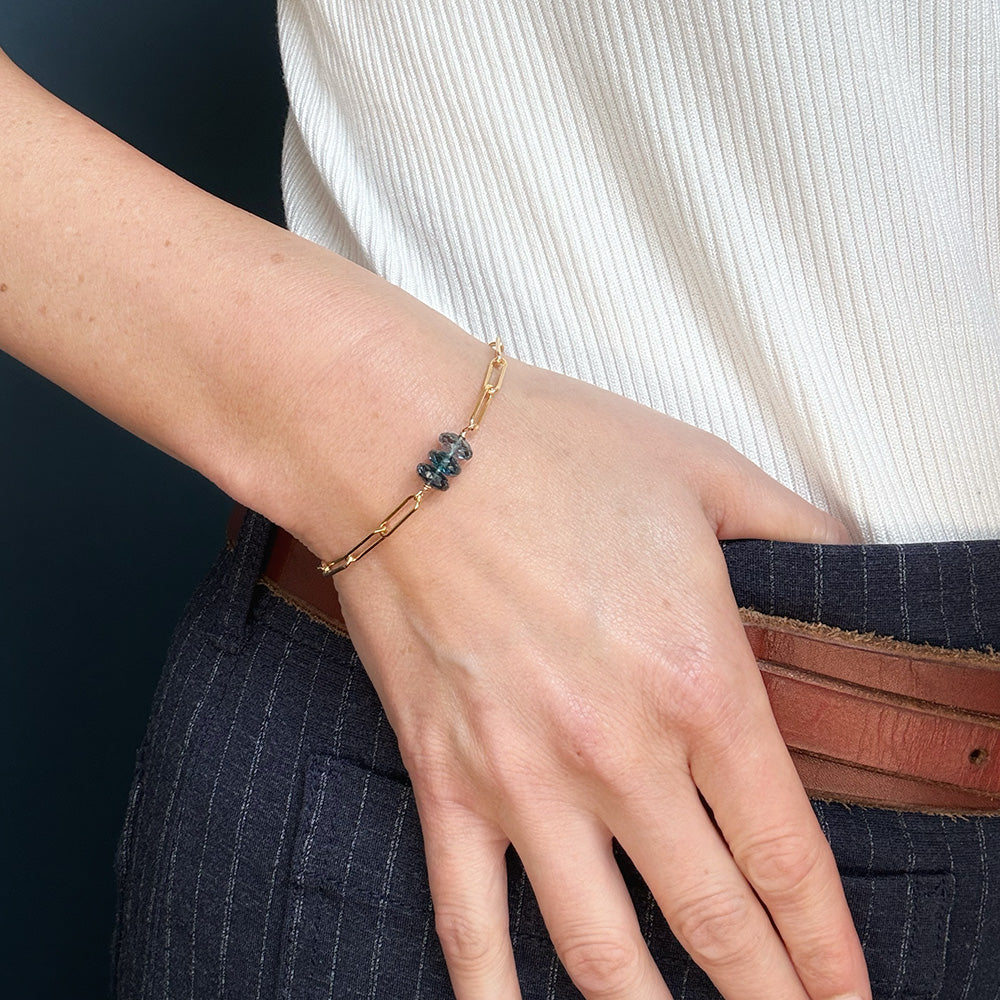 A close-up of a person's hand wearing a delicate gold bracelet with a small cluster of London Blue Topaz gemstones, styled with a white ribbed top and dark pinstriped trousers.
