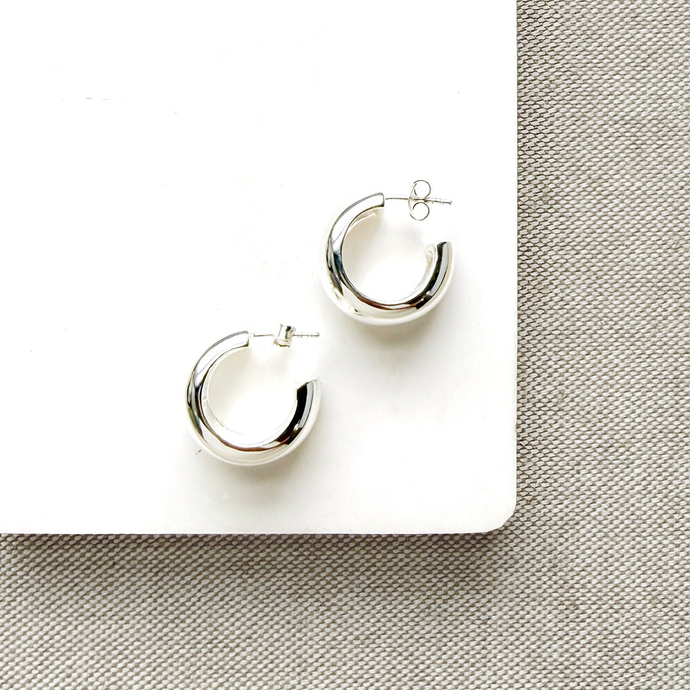 A pair of polished silver crescent-shaped hoop earrings displayed on a white square card over a textured gray fabric background.