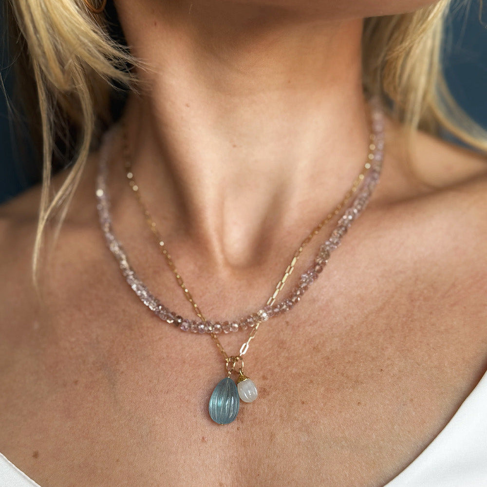 A close-up of a model wearing an ametrine gemstone necklace with a chain and charms made of carved aquamarine and carved white chalcedony.