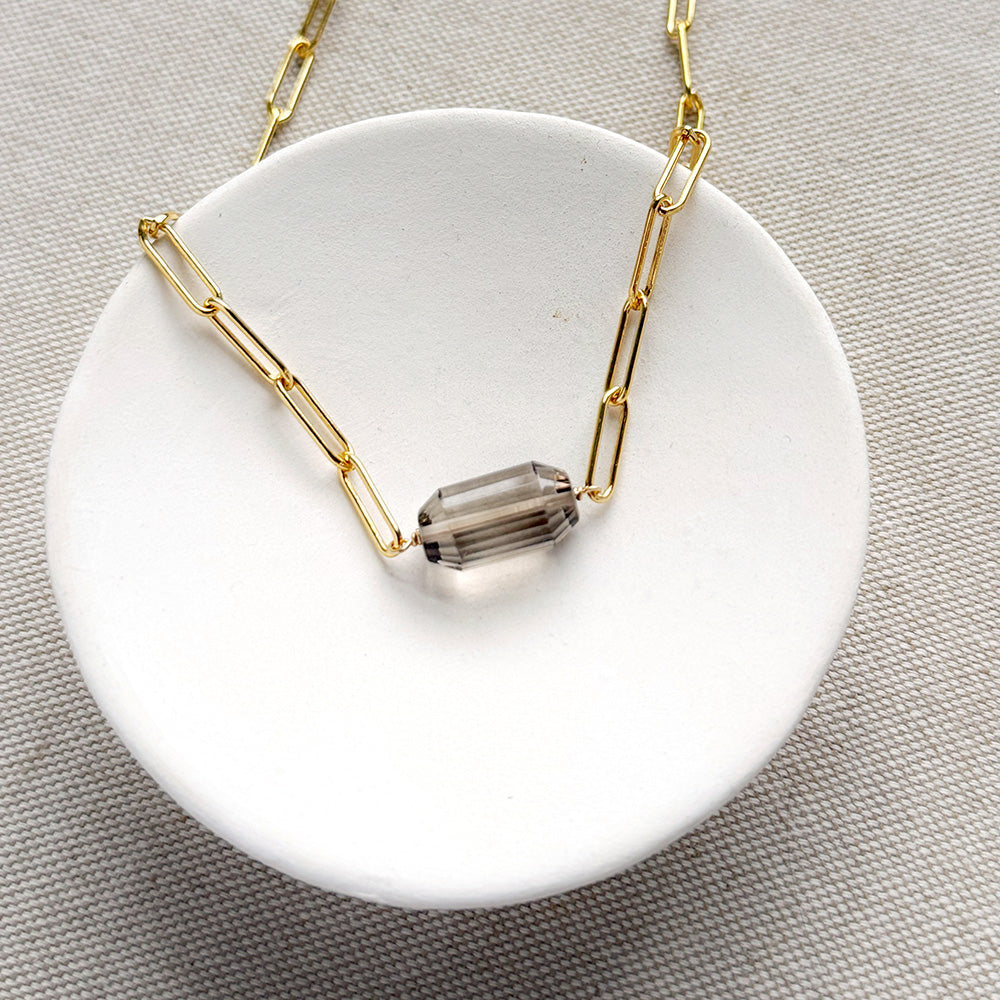 A gold paperclip chain necklace with a center set smokey quartz, displayed on a white card over a textured gray fabric background.