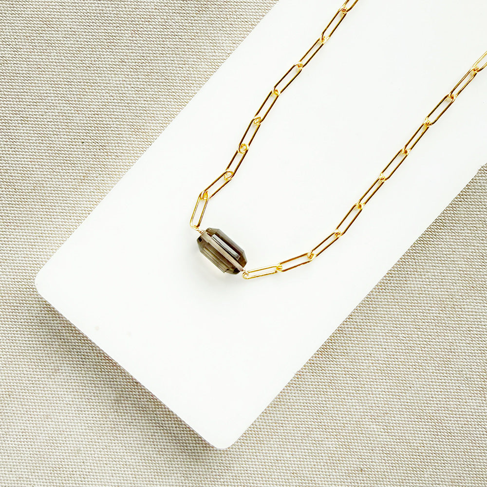 A gold paperclip chain necklace with a center set smokey quartz, displayed on a white card over a textured gray fabric background.