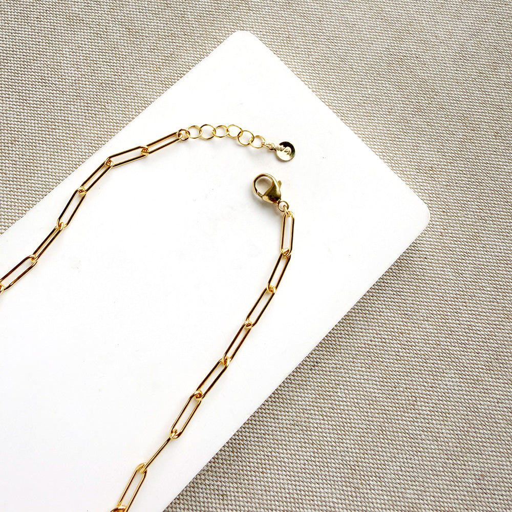 A gold paperclip chain necklace with a lobster clasp and extender, laid on a white rectangular card over a textured beige fabric background.