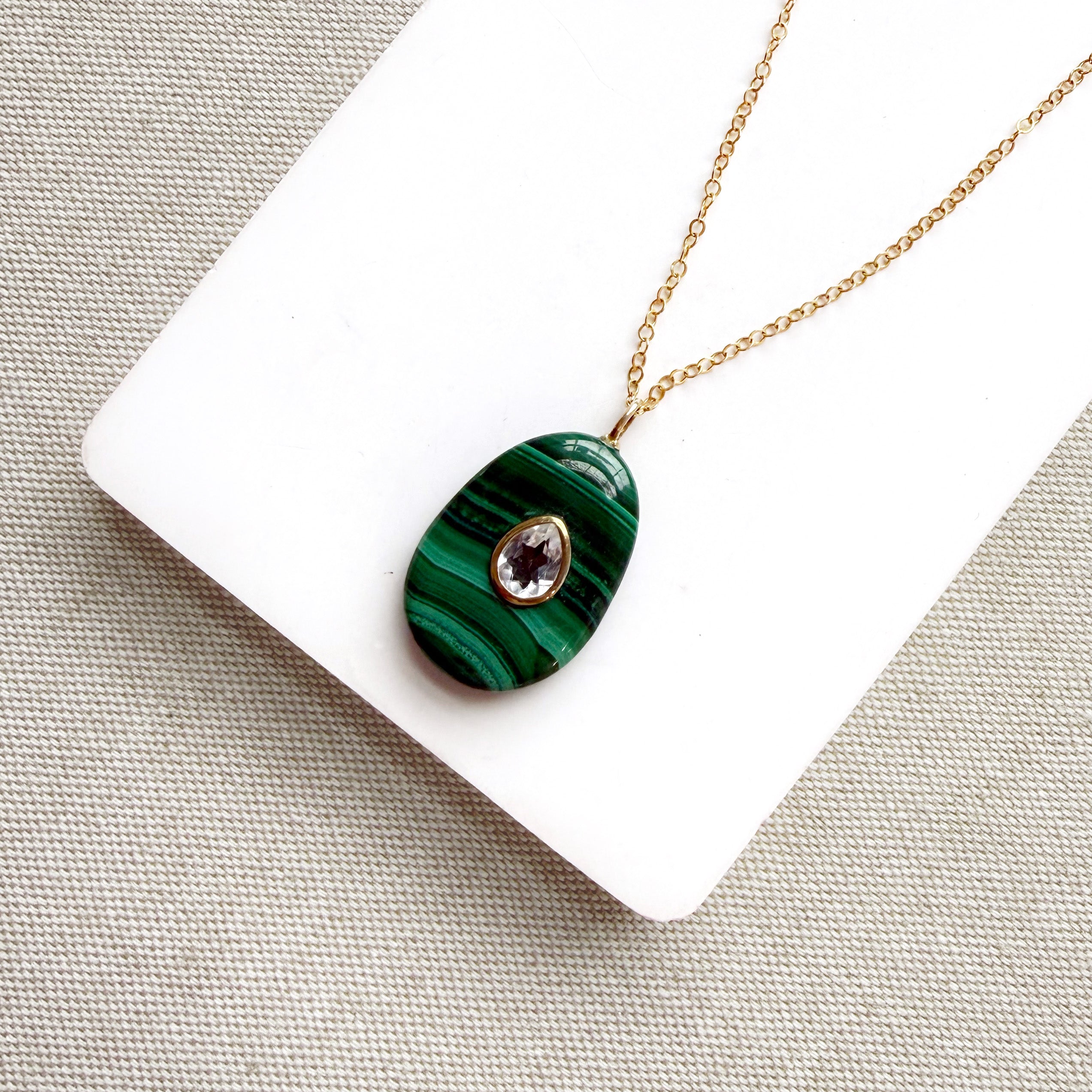 A gold chain necklace with an oval green malachite pendant featuring a small teardrop-shaped purple gemstone in the center, displayed on a white card over a textured beige fabric background.
