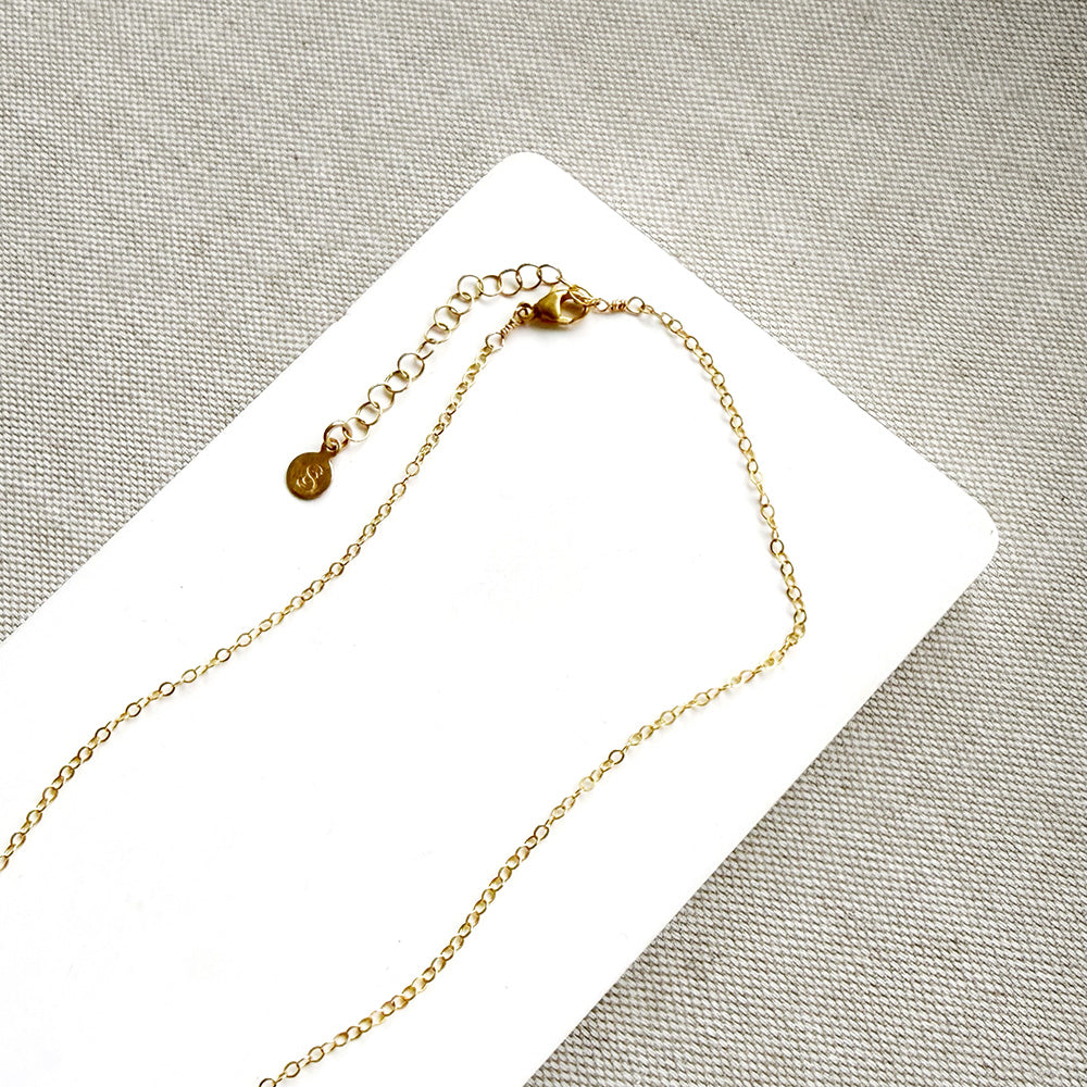 A delicate gold chain necklace with an adjustable lobster clasp and a small round engraved charm, displayed on a white card over a textured gray fabric background.
