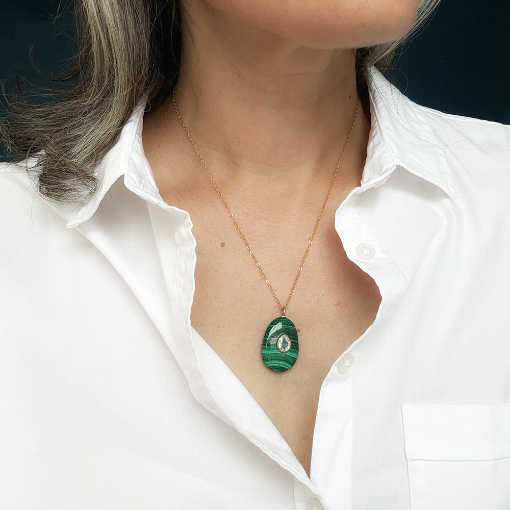 A close-up of a person wearing a delicate gold chain necklace with an oval green stone pendant featuring a small white inlay, styled with a white button-down shirt.