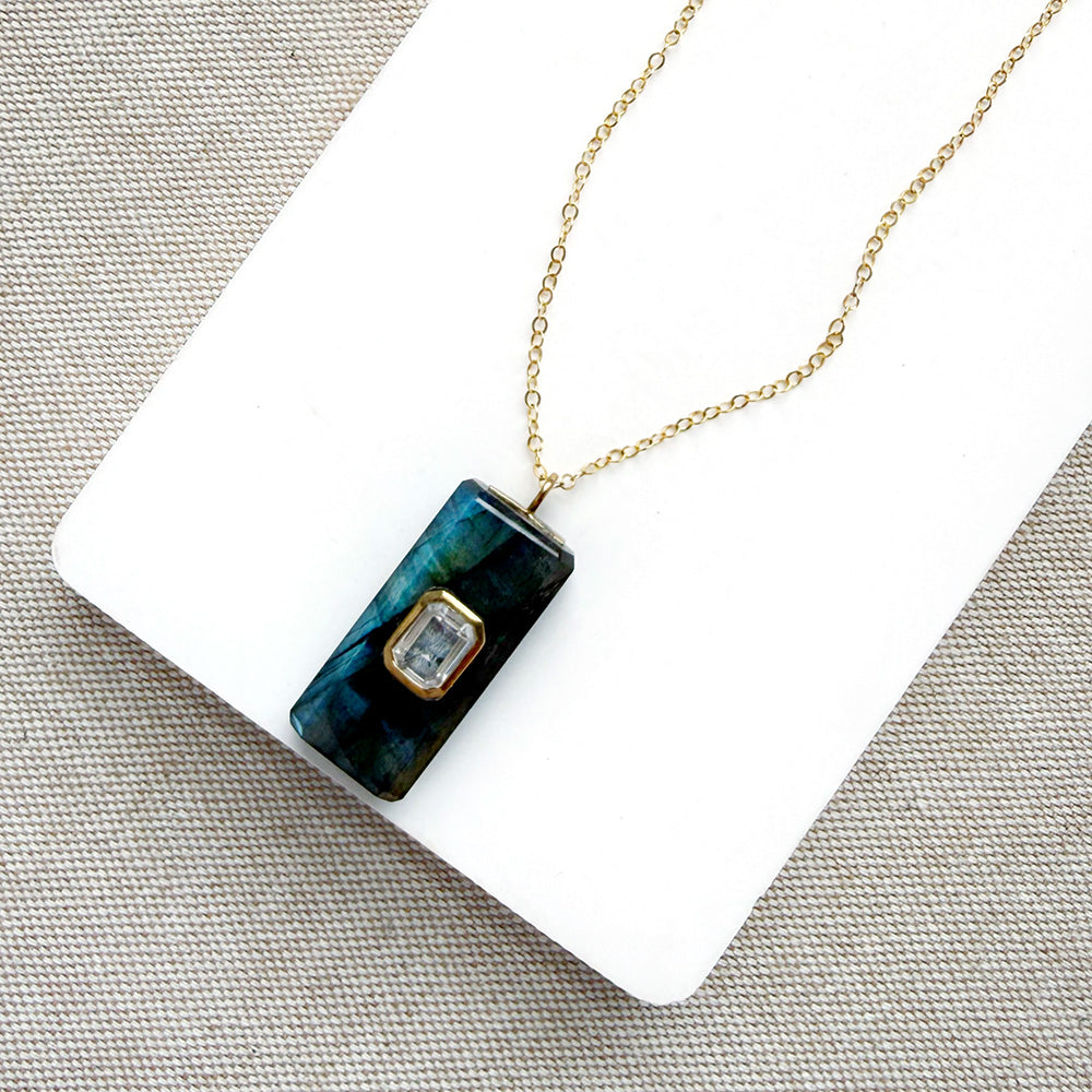 A gold chain necklace with a rectangular dark teal and green stone pendant featuring a small gold-set clear gemstone, displayed on a white card over a textured gray fabric background.