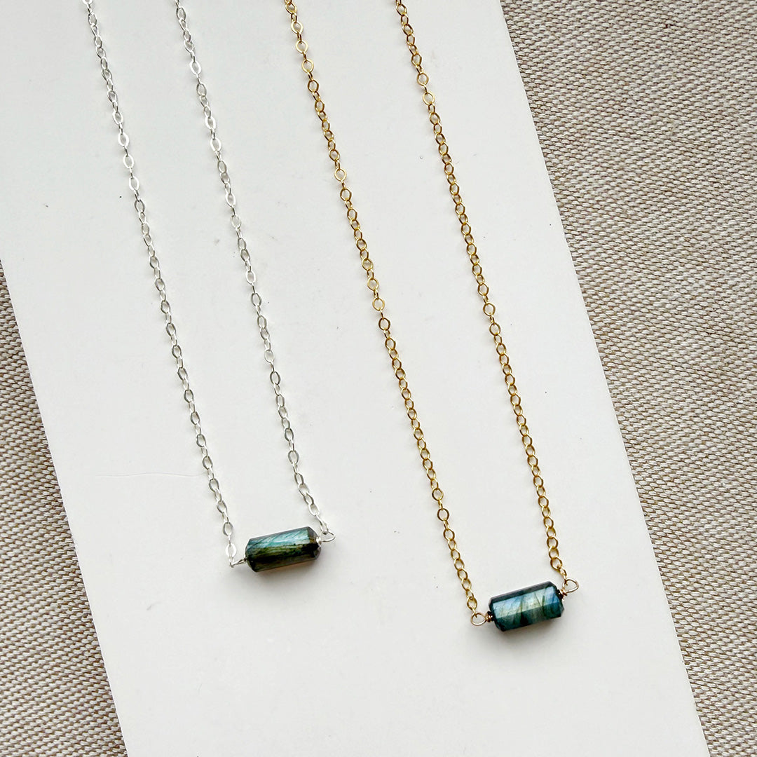 Two necklaces, one silver and one gold, with a single labradorite gemstone on a white dish against a textured surface