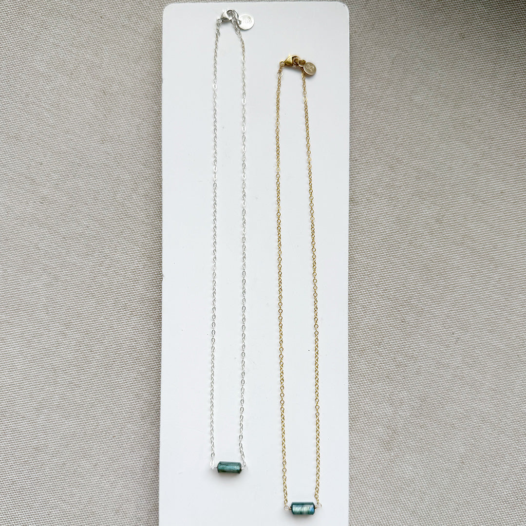 Two necklaces, one silver and one gold, with a single labradorite gemstone on a white dish against a textured surface