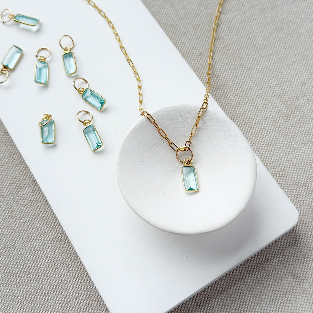 Gold-tone necklace with emerald cut aquamarine charm, displayed on a white dish and matching white rectangular board over a neutral fabric background.