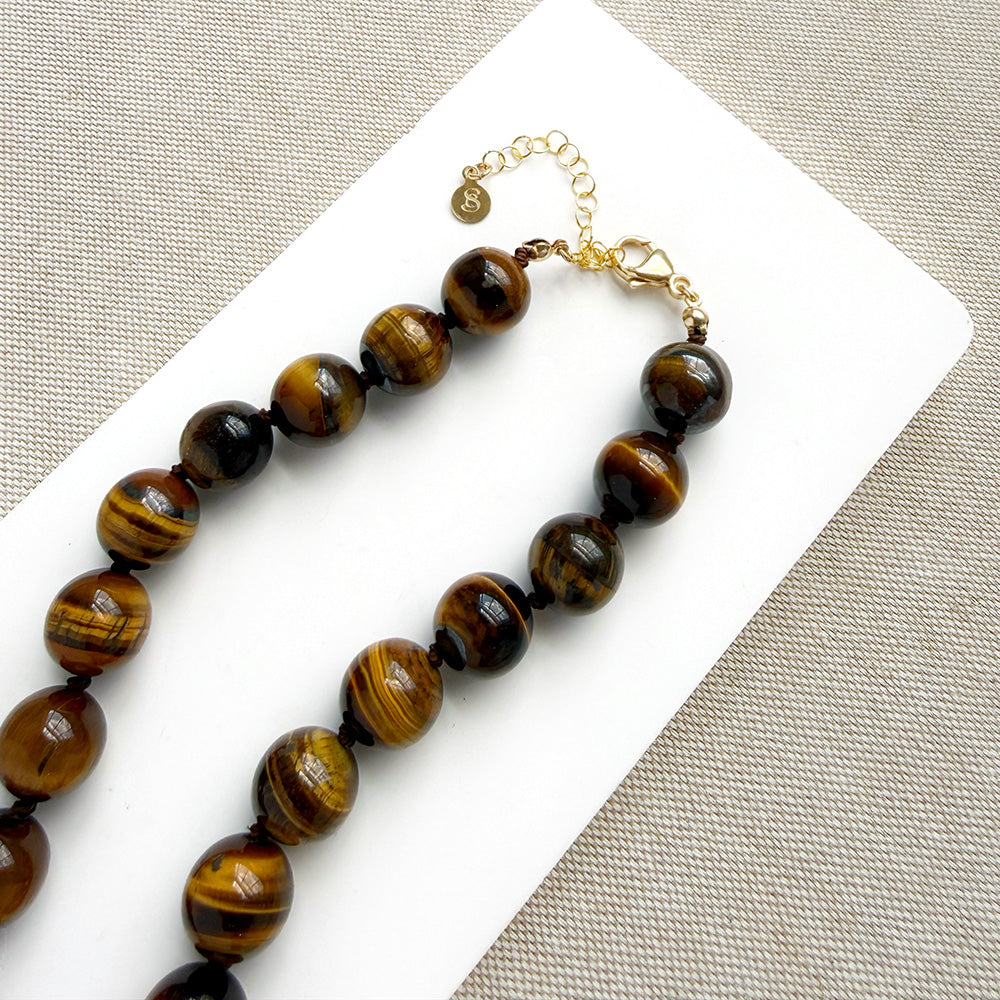 A beaded necklace made of polished tiger's eye beads in brown and gold tones, draped over a small white dish on a white rectangular board over a textured beige fabric background.