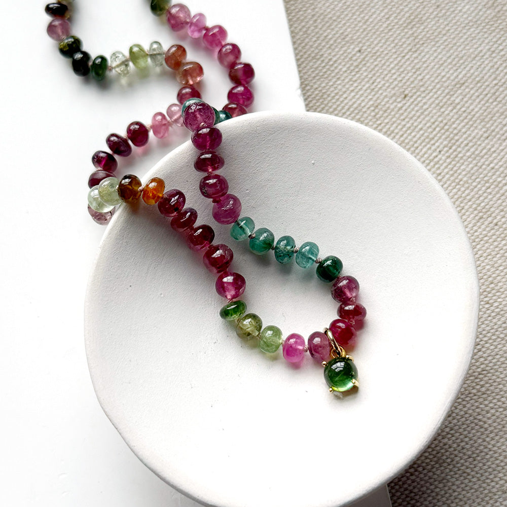 Smooth Watermelon Tourmaline beads in shades of pink, red, green, blue, and orange, featuring a small green tourmaline charm with gold detailing, displayed on a white ceramic dish.