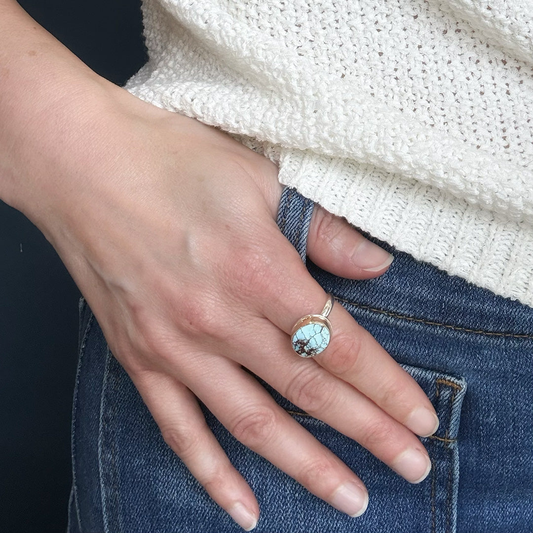 Hand wearing a one of a kind mixed metal Golden Hills Turquoise ring on a dark background
