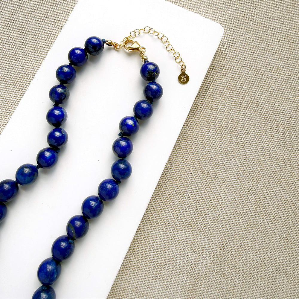 A beaded necklace made of polished dark blue lapis lazuli stones with a gold clasp and extender chain, displayed on a white rectangular card over a beige woven fabric background.