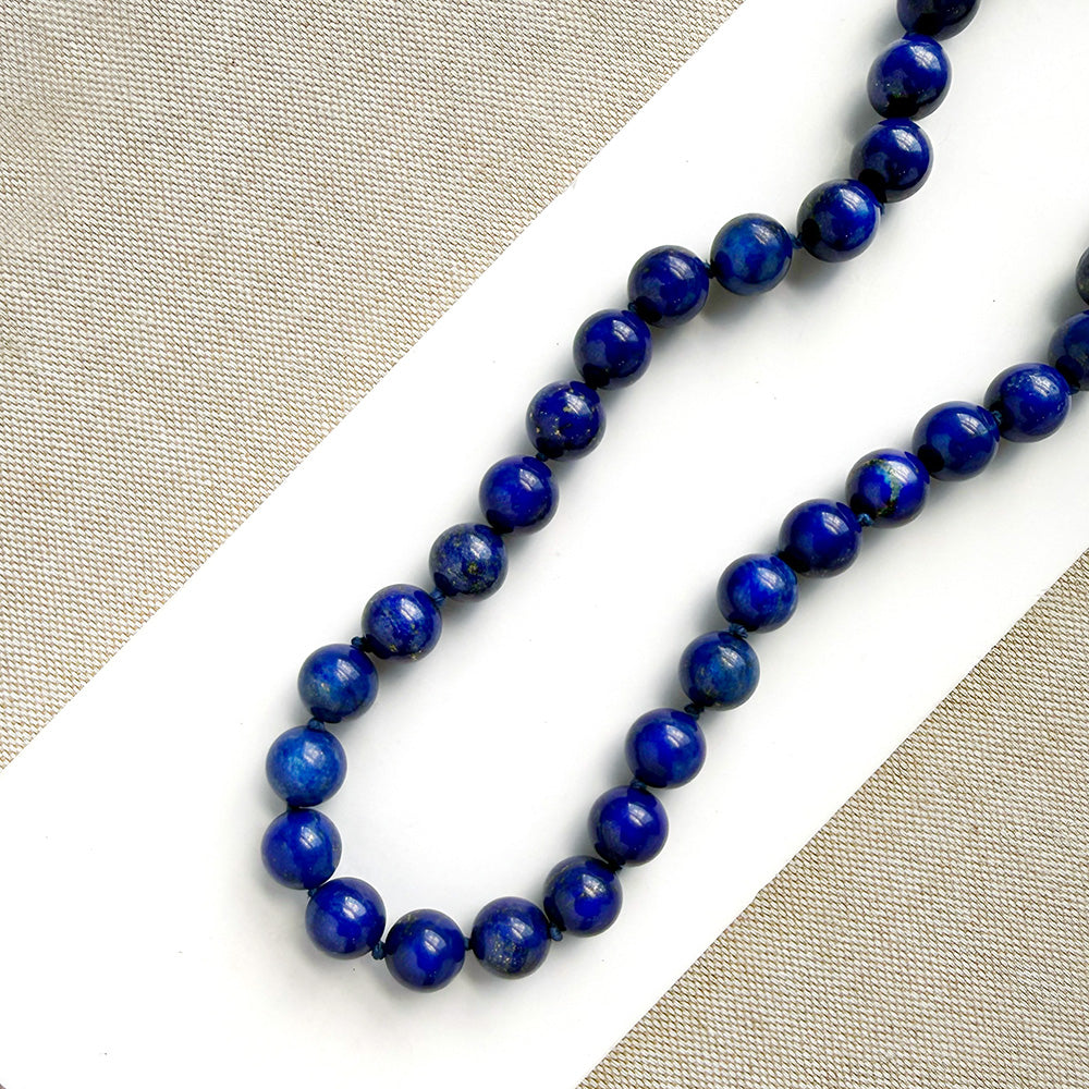 A strand of polished dark blue lapis lazuli ball beads arranged diagonally on a white surface with a beige textured fabric background.