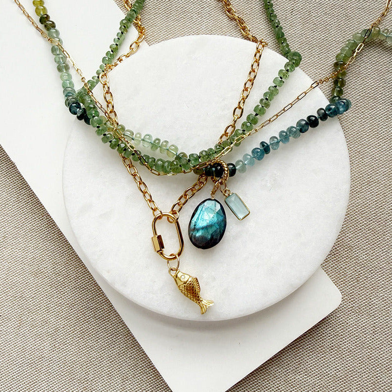 A layered gold-tone necklace set displayed on a white circular stand over a beige fabric background. The necklace features green and blue-green beaded strands with a teardrop-shaped blue-green stone pendant and a small gold fish charm.