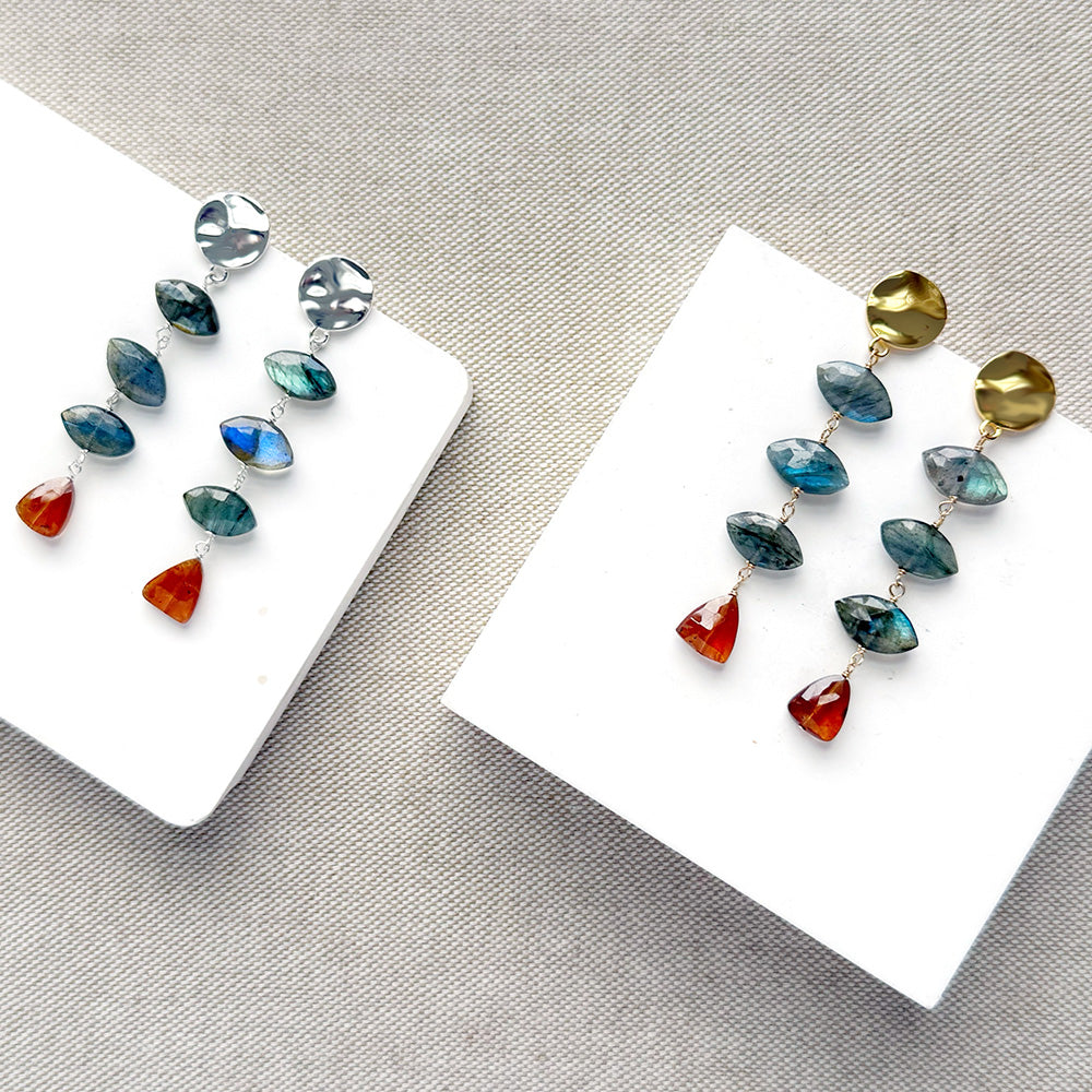 One pair of gold and one pair of silver dangle earrings with marquis cut labradorite gems and a triangular hessian garnet, displayed on a white square card over a textured beige fabric background.