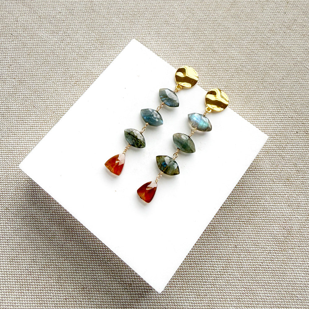 A pair of gold dangle earrings with marquis cut labradorite gems and a triangular hessian garnet, displayed on a white square card over a textured beige fabric background.
