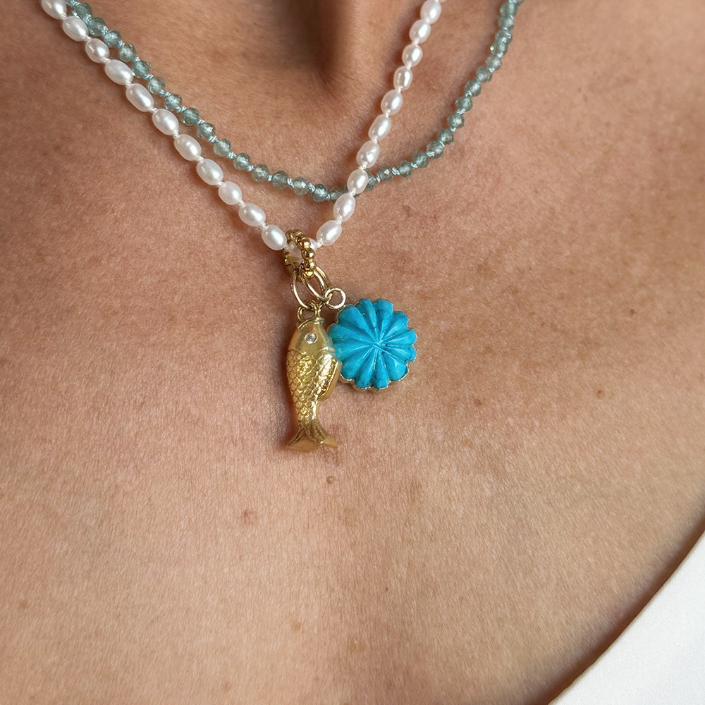 A close-up of a model wearing layered necklaces with white pearls and a light blue-green gemstones. The necklace features a gold-tone fish charm and a small howlite flower pendant.