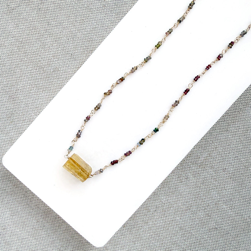 A multi-color sapphire necklace with a central imperial topaz pendant, displayed on a white background over a gray textured fabric background.