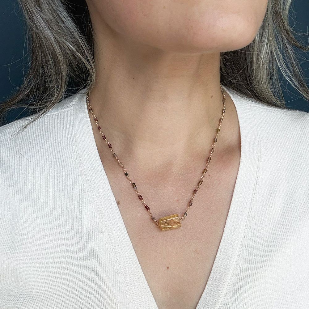 A close-up of a person wearing a multi-color sapphire necklace with a central imperial topaz pendant, paired with a white V-neck top.