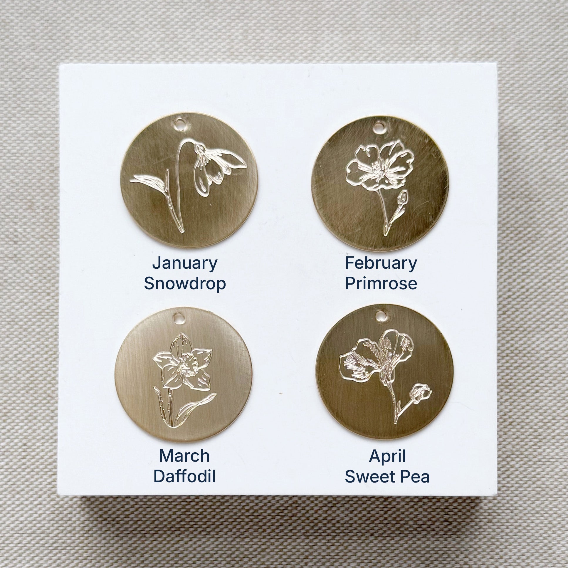 Four gold floral charms on a white card with month and flower name labels, on a textured beige background.
