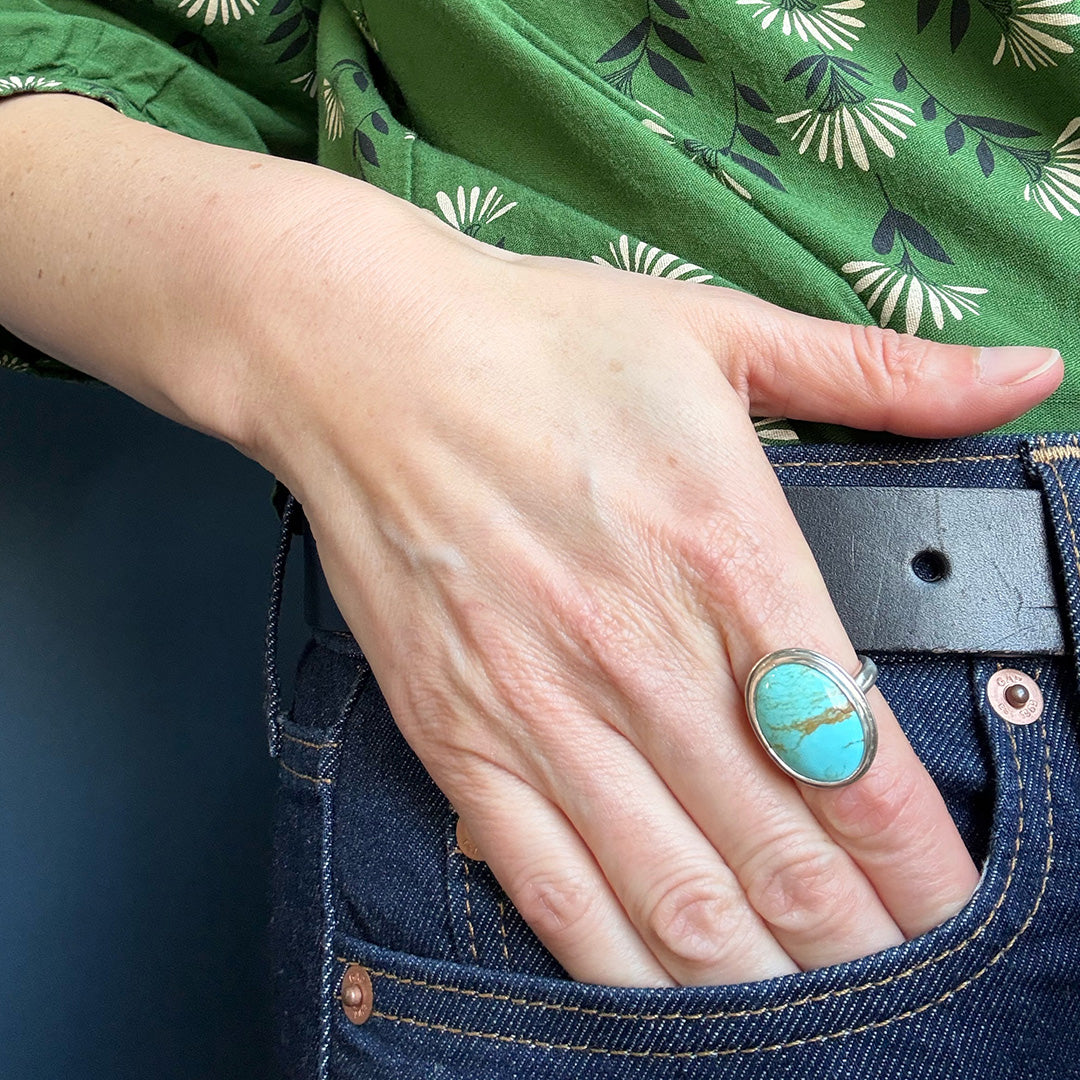 Hand wearing a turquoise ring with a green floral-patterned shirt and blue jeans.