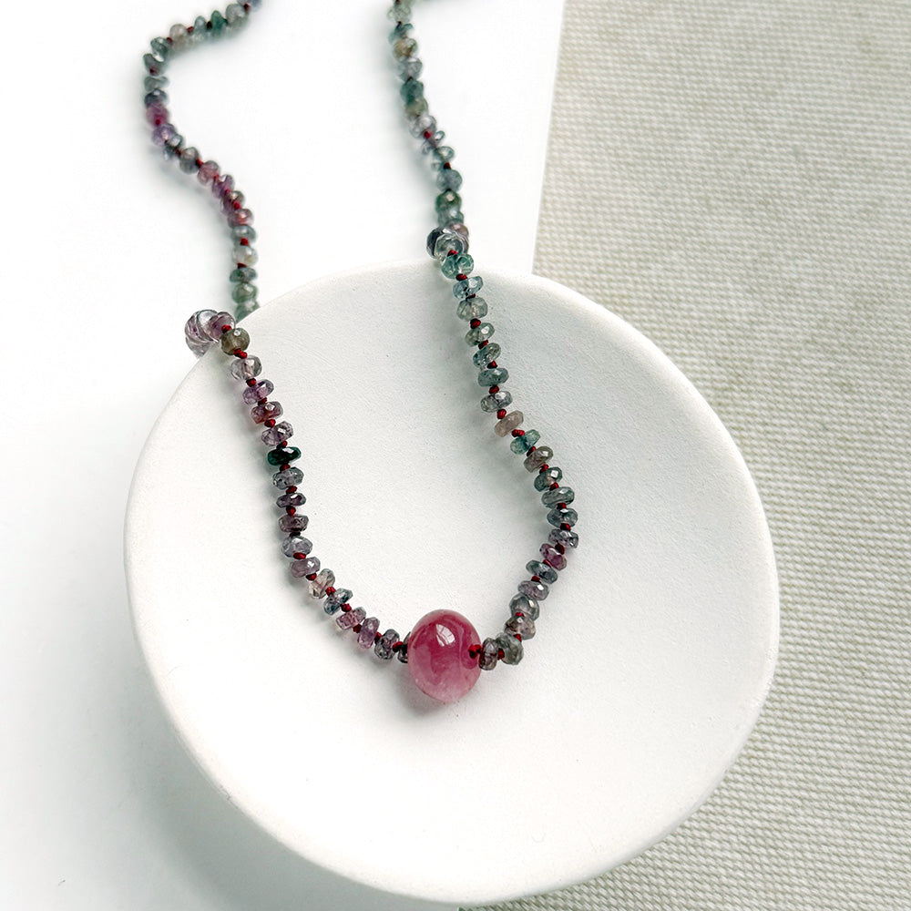 A necklace with small multicolored sapphire beads and a prominent oval pink sapphire at the center, displayed on a white ceramic dish against a light textured background.