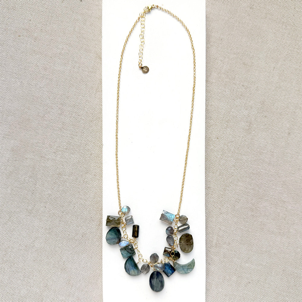 A gold chain necklace with multiple faceted labradorite gemstone charms in shades of blue, green, gray, and black, arranged on a white card over a beige woven fabric background.
