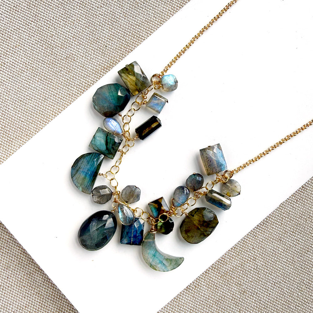 A gold chain necklace with multiple faceted labradorite gemstone charms in shades of blue, green, gray, and black, arranged on a white card over a beige woven fabric background.