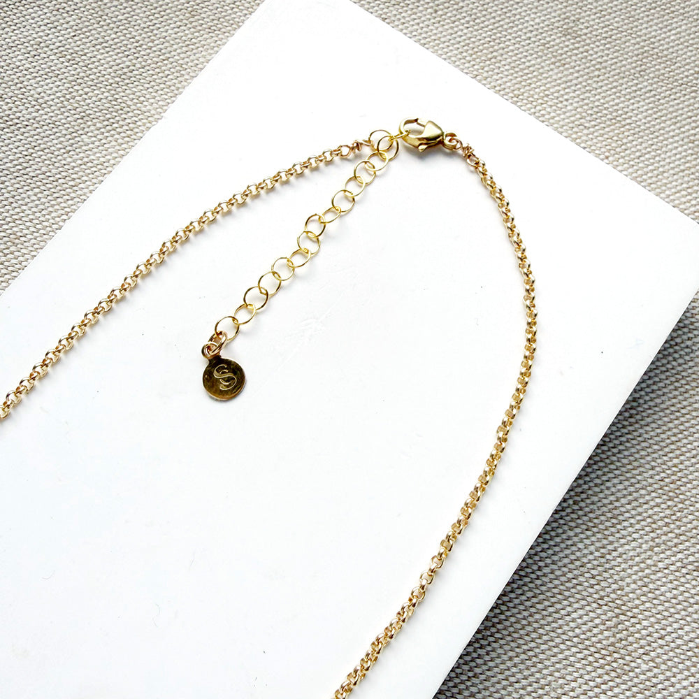 Close-up of a gold chain necklace with a lobster clasp, extension chain, and a small round engraved disc charm, laid on a white rectangular surface over a textured gray fabric background.