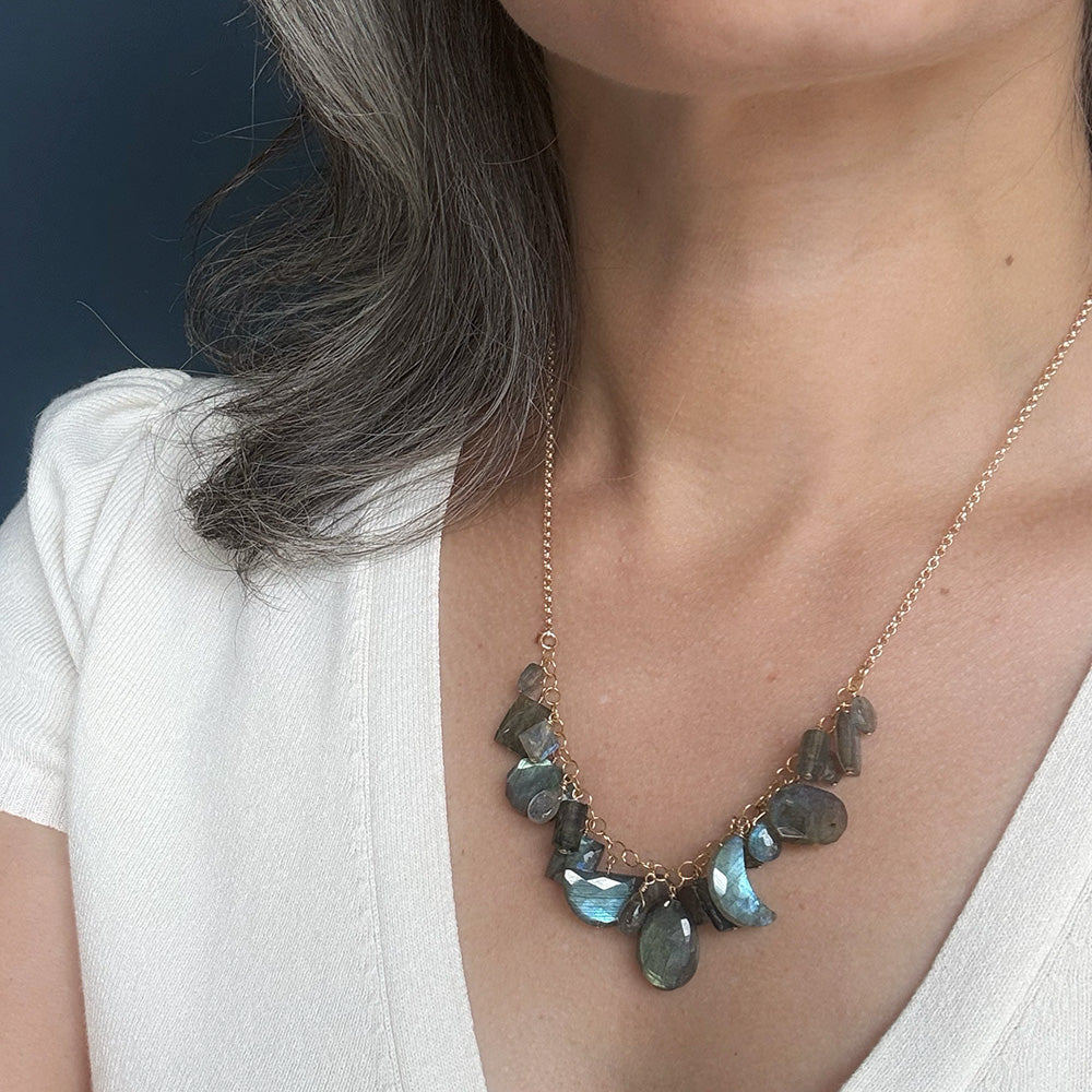 A close-up of a person wearing a gold chain necklace with faceted gray-blue labradorite stones, styled with a white top against a dark blue background.