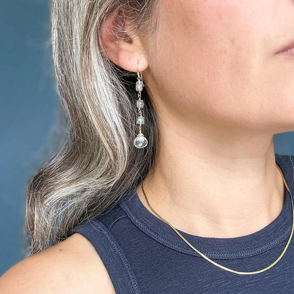 A close-up of a woman wearing a delicate gold drop earring with small clear faceted beads and a larger clear teardrop charm. The earring is shown on her ear, with her gray hair and navy blue top visible against a soft blue-gray background.