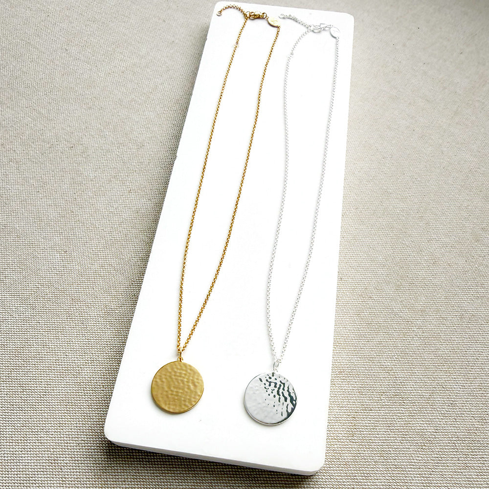 Two round pendant necklaces displayed on a white rectangular card against a neutral beige fabric background. One necklace is gold with a hammered disc pendant, and the other is silver with a hammered disc pendant.