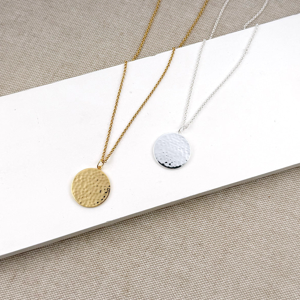 Two minimalist hammered round pendant necklaces displayed on a white rectangular block over a beige textured fabric background. One necklace has a gold chain and a gold pendant, and the other has a silver chain and a silver pendant.